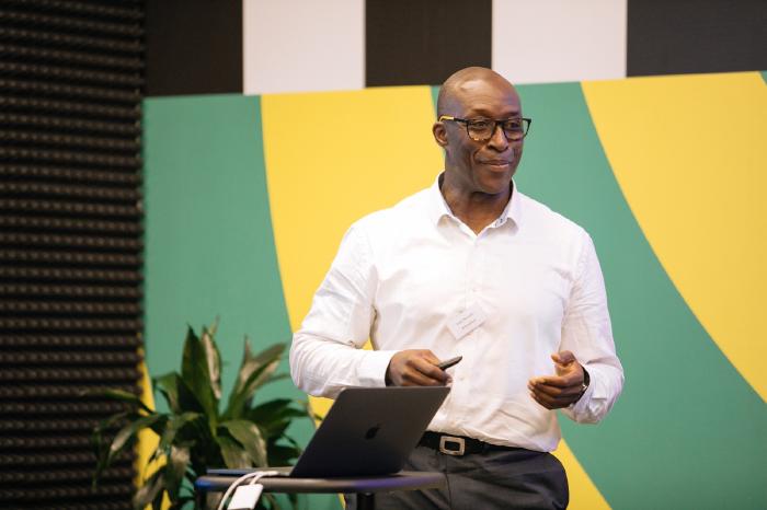 Fredi Nonyelu presenting with a laptop at a technology conference, standing in front of a colourful backdrop