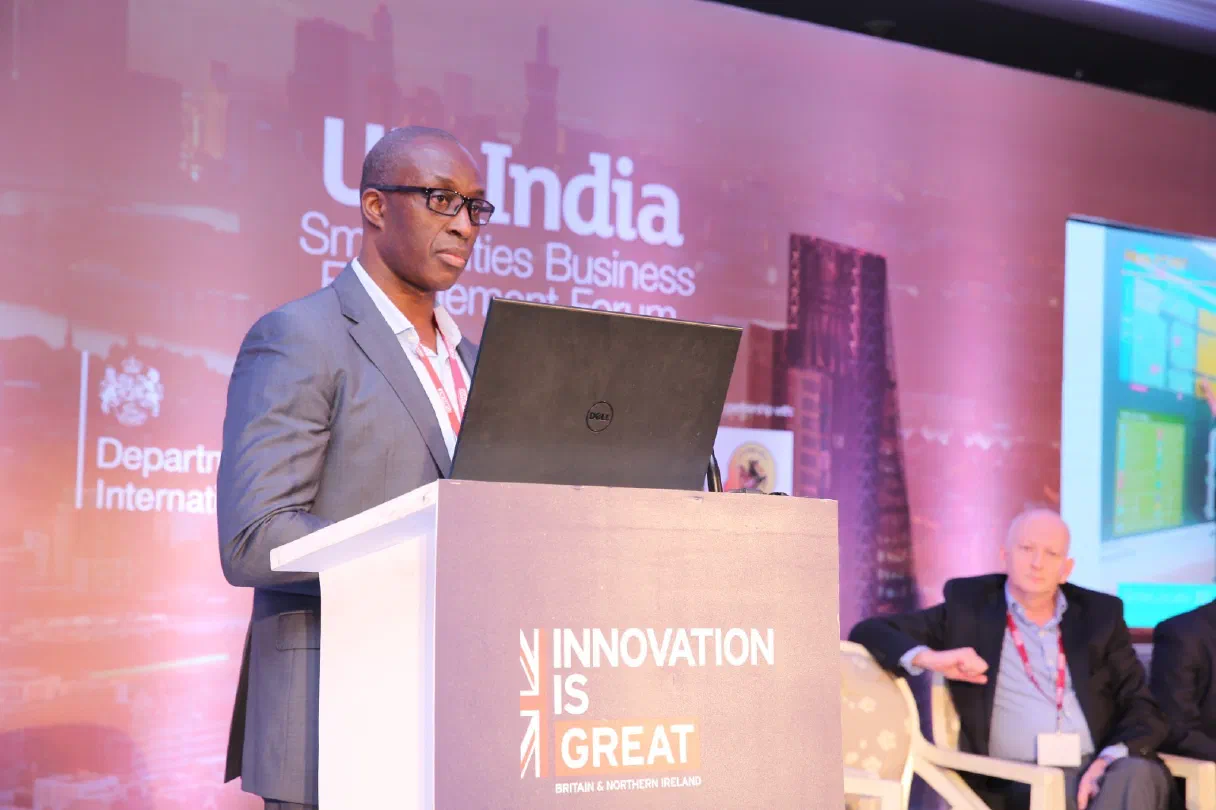 Fredi Nonyelu presenting with a laptop at a technology conference, standing in front of a colourful backdrop